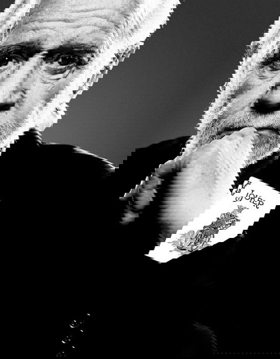 Portrait of George Carlin with a joker playing card coming out of his shirt sleeve